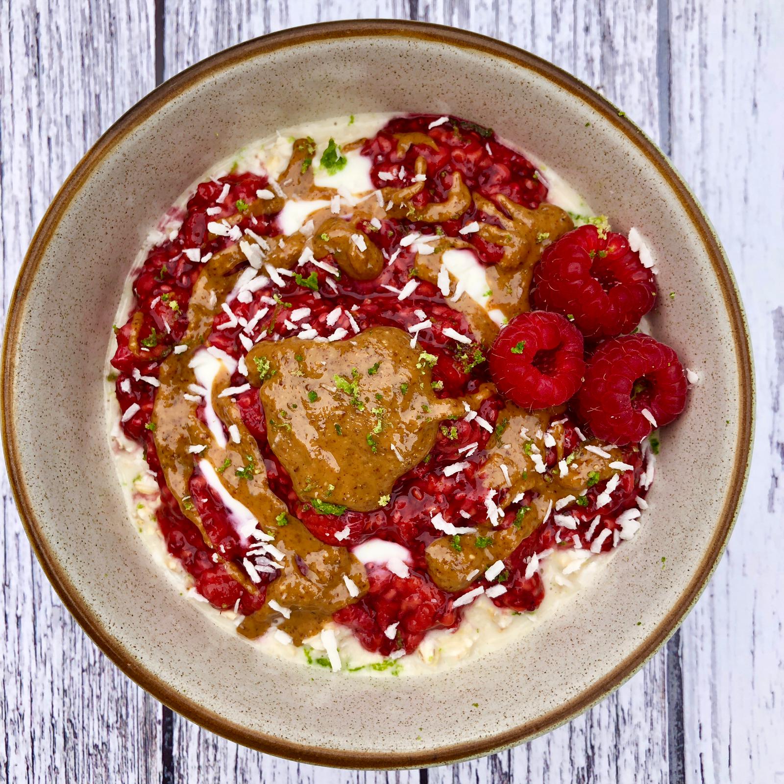 Bowl of overnight oats topped with fresh raspberries, raspberry chia jam, nut butter, lime zest and coconut