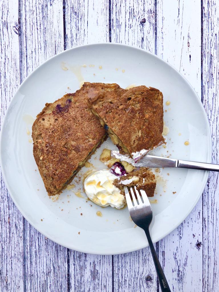 Banana and blueberry cinnamon french toast on a plate with a knife and fork, served with yoghurt and honey