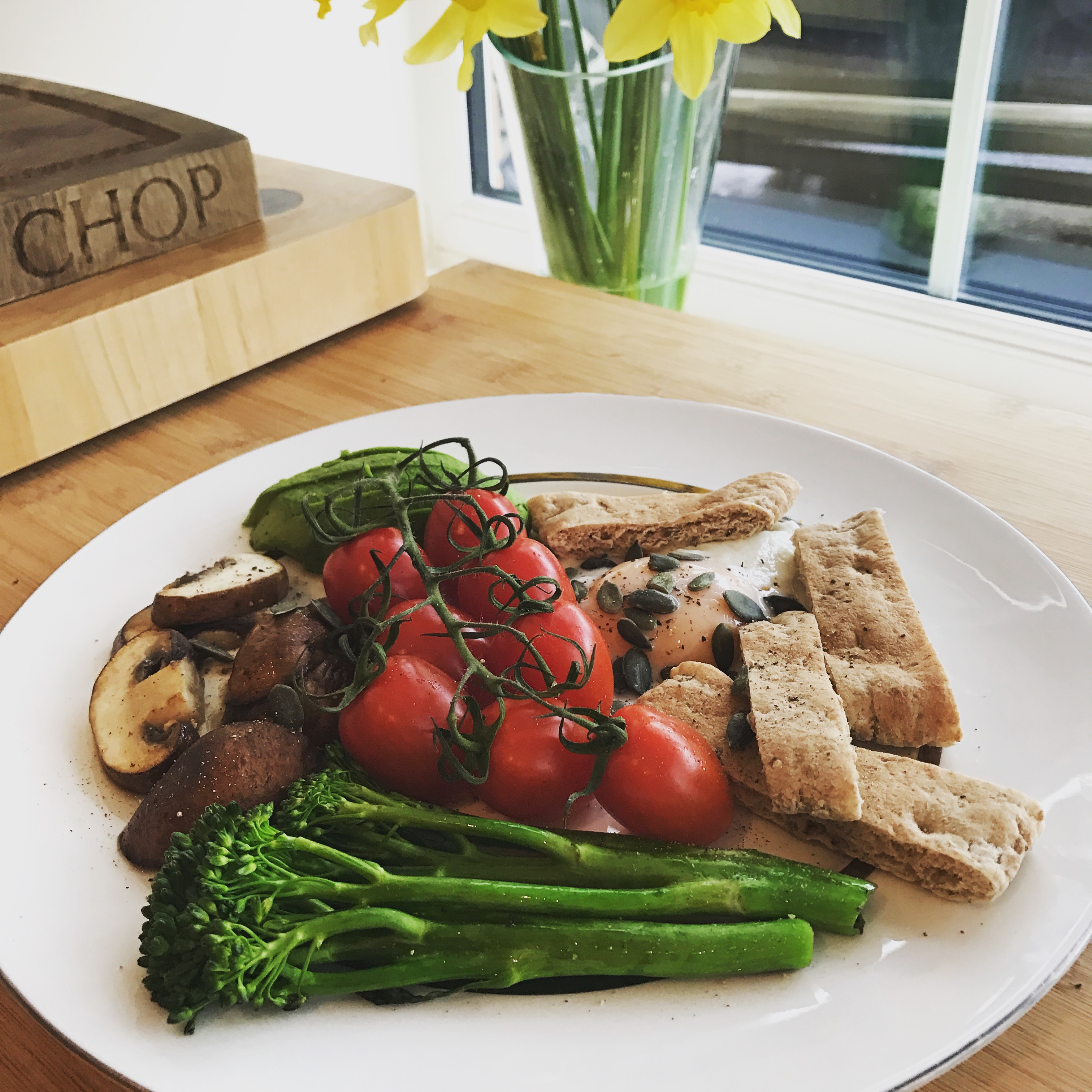 Plate with fried egg, sliced avocado, sliced mushrooms, vine tomatoes, tenderstem broccoli, sliced pitta and sunflower seeds on a table by the window