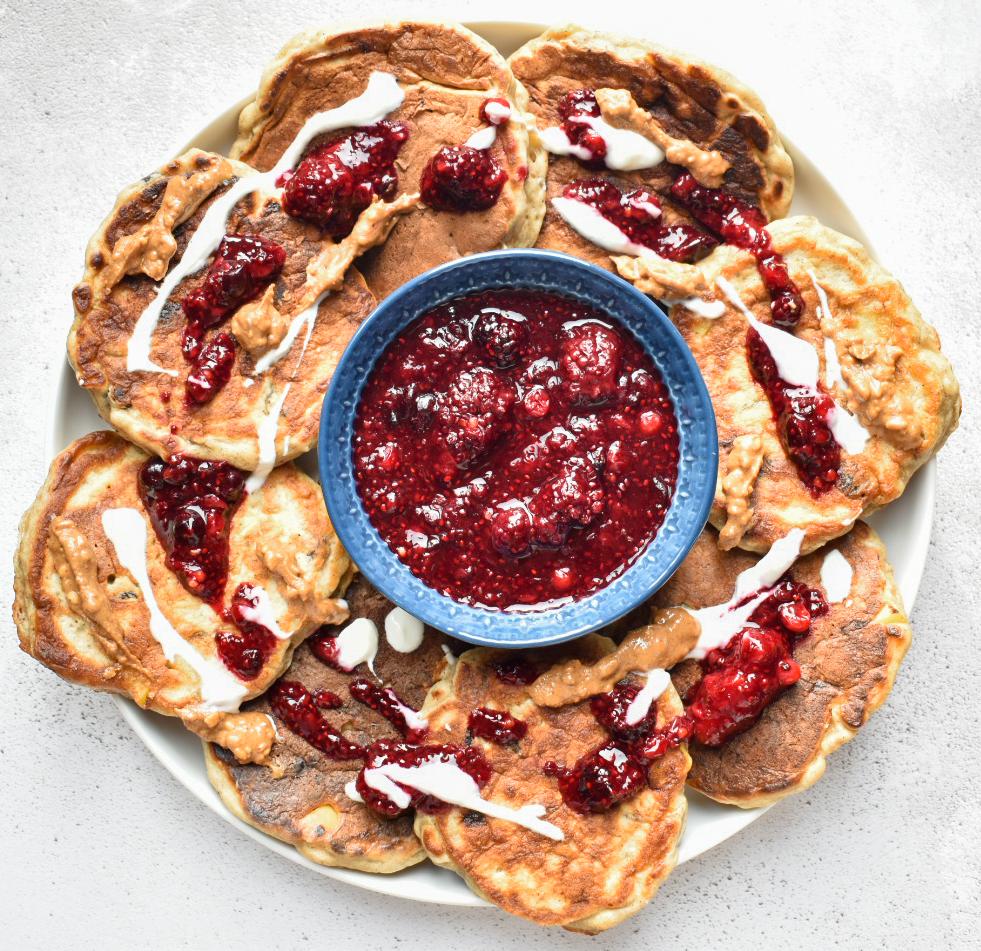 Bowl of berry compote surrounded by small apple and raisin pancakes topped with berry compote, yoghurt and nut butter