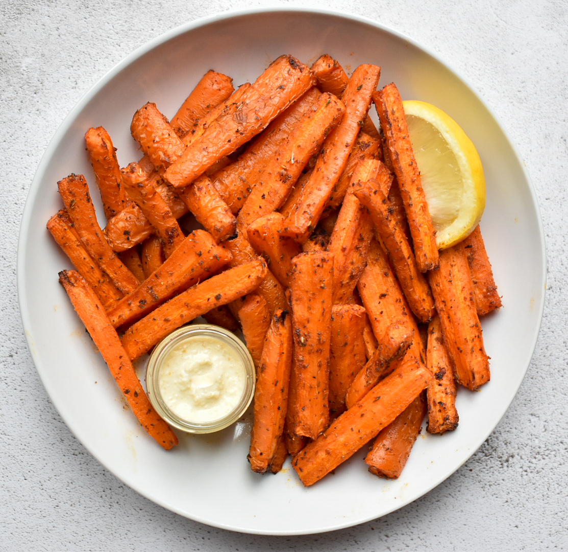 Baked spicy carrot sticks served on a plate with a jar of hummus and a wedge of lemon