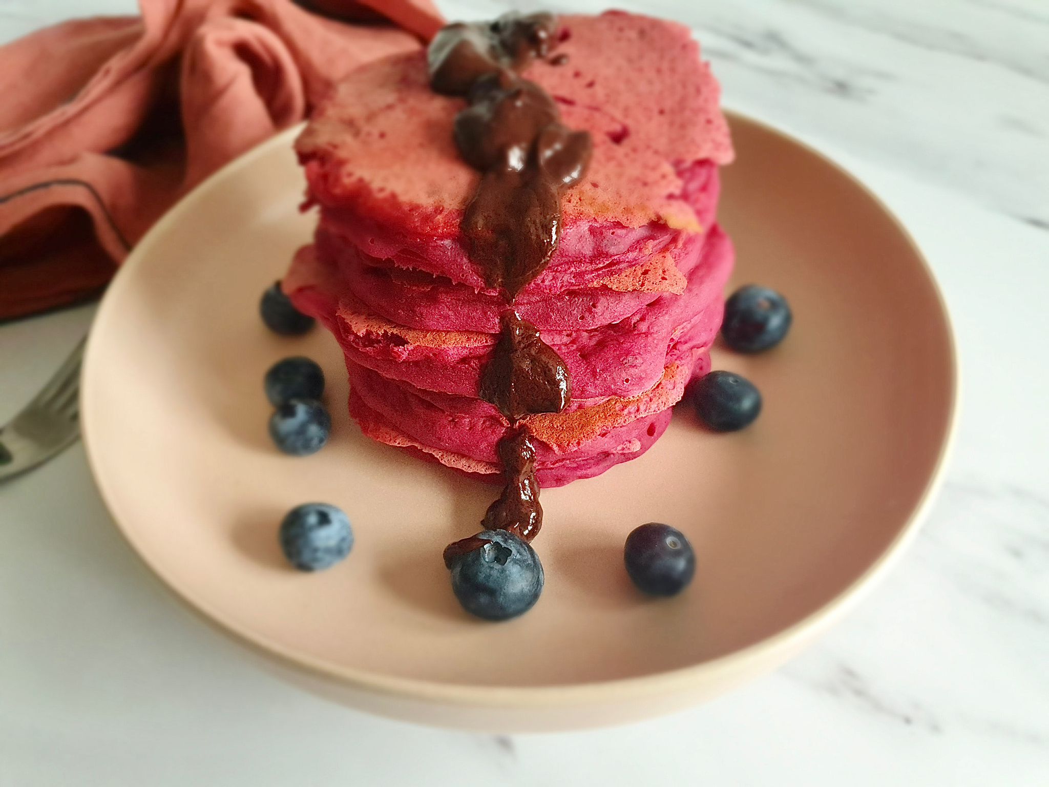 Stack of beetroot pancakes on a plate topped with melted dark chocolate and blueberries