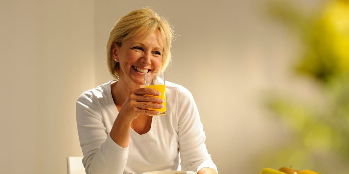 1 middle aged woman sat at a kitchen table holding a glass of orange juice and smiling