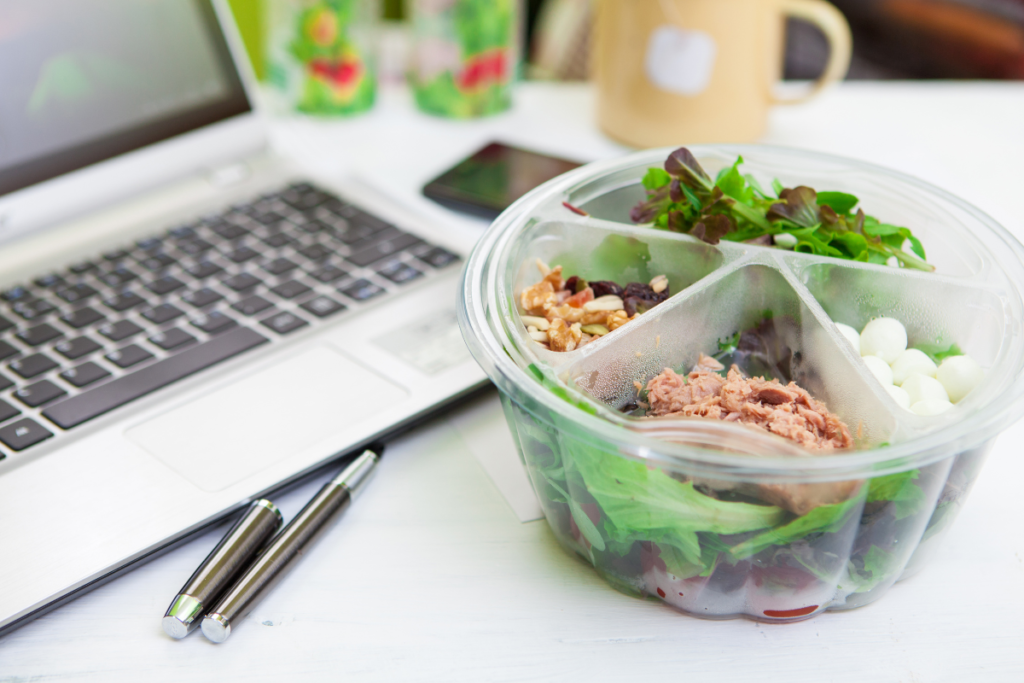 a salad lunch box on a desk next to a laptop, phone and pens