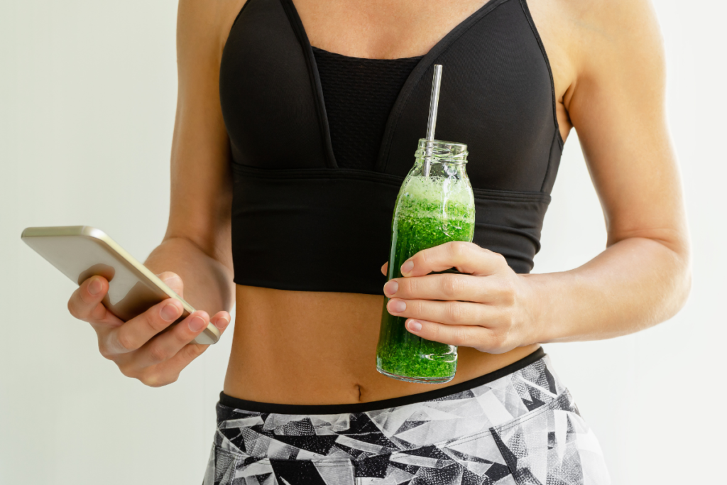 young woman wearing gym gear holding a green smoothie and checking mobile phone