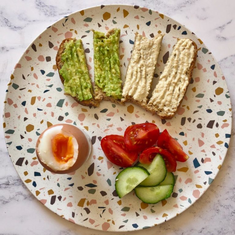 Plate with a piece of toast cut into 4, half with mashed avocado and half with hummus, served with a boiled egg, sliced tomato and cucumber