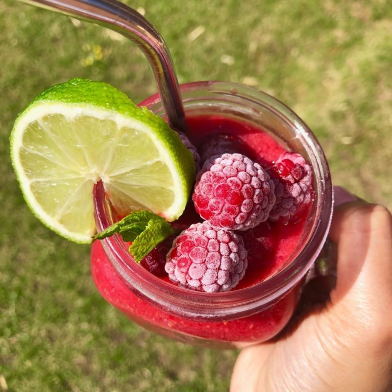 Glass of fruity slush garnished with frozen raspberries, mint leaves and lime slice